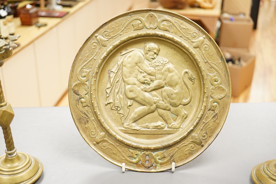 A pair of Pugin style ‘gothic’ brass candlesticks - 21cm high and a circular brass disc depicting Hercules and The Nemean Lion - 25cm in diameter